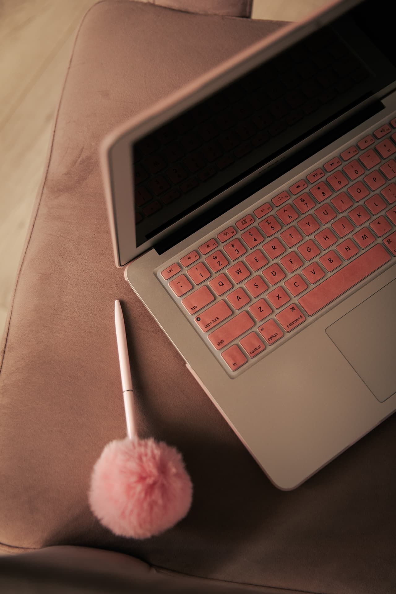 A laptop with pink keyboard cover and a pom pom pen
